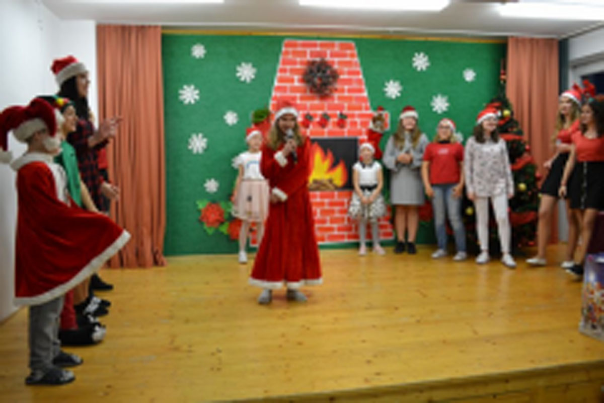 Uczniowie siedzą na krzesłach. Na głowach mają czerwone czapki Mikołaja. Oglądają występ na scenie. Mikołaj siedzi w fotelu na scenie i rozdaje prezenty.