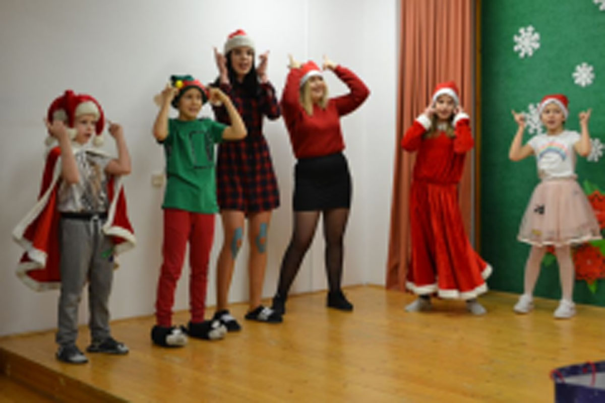 Uczniowie siedzą na krzesłach. Na głowach mają czerwone czapki Mikołaja. Oglądają występ na scenie. Mikołaj siedzi w fotelu na scenie i rozdaje prezenty.