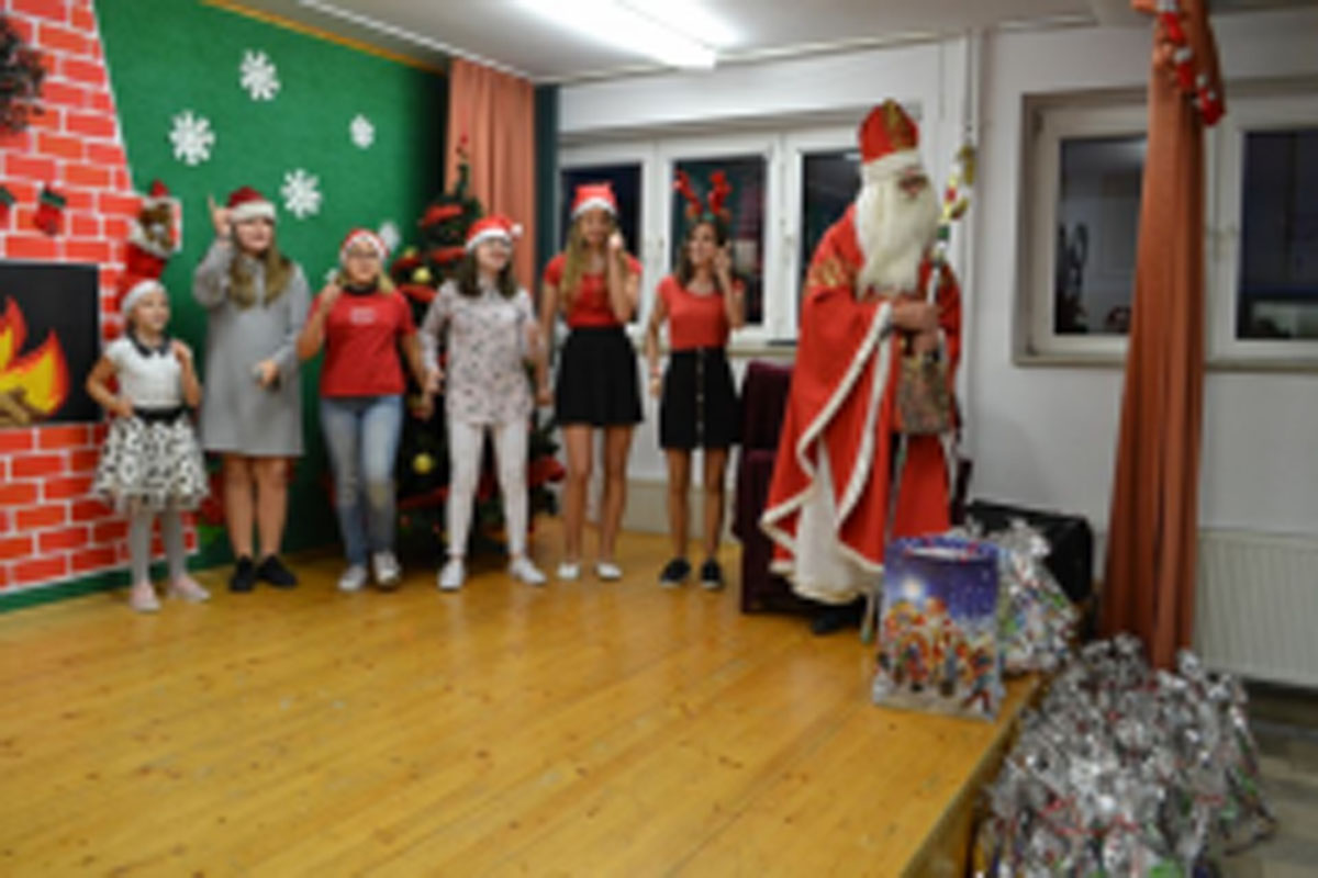 Uczniowie siedzą na krzesłach. Na głowach mają czerwone czapki Mikołaja. Oglądają występ na scenie. Mikołaj siedzi w fotelu na scenie i rozdaje prezenty.