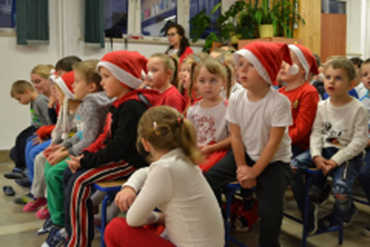 Uczniowie siedzą na krzesłach. Na głowach mają czerwone czapki Mikołaja. Oglądają występ na scenie. Mikołaj siedzi w fotelu na scenie i rozdaje prezenty.