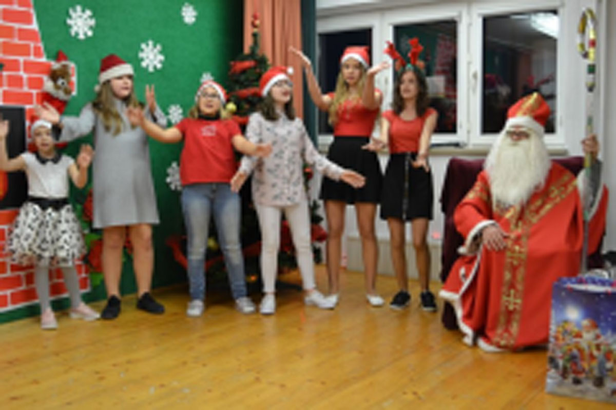 Uczniowie siedzą na krzesłach. Na głowach mają czerwone czapki Mikołaja. Oglądają występ na scenie. Mikołaj siedzi w fotelu na scenie i rozdaje prezenty.