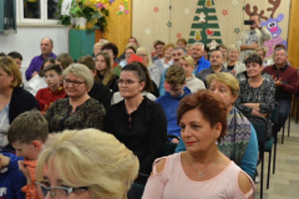 Uczniowie siedzą na krzesłach. Na głowach mają czerwone czapki Mikołaja. Oglądają występ na scenie. Mikołaj siedzi w fotelu na scenie i rozdaje prezenty.