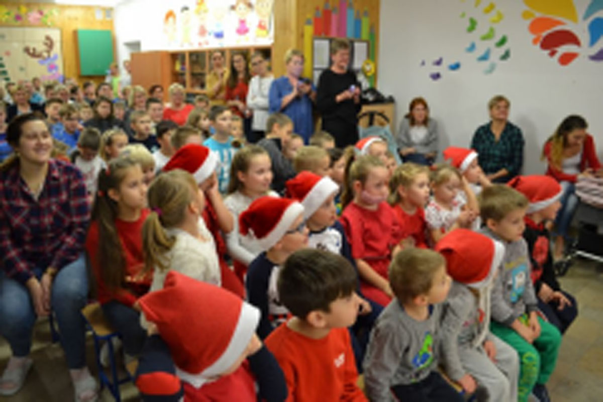 Uczniowie siedzą na krzesłach. Na głowach mają czerwone czapki Mikołaja. Oglądają występ na scenie. Mikołaj siedzi w fotelu na scenie i rozdaje prezenty.