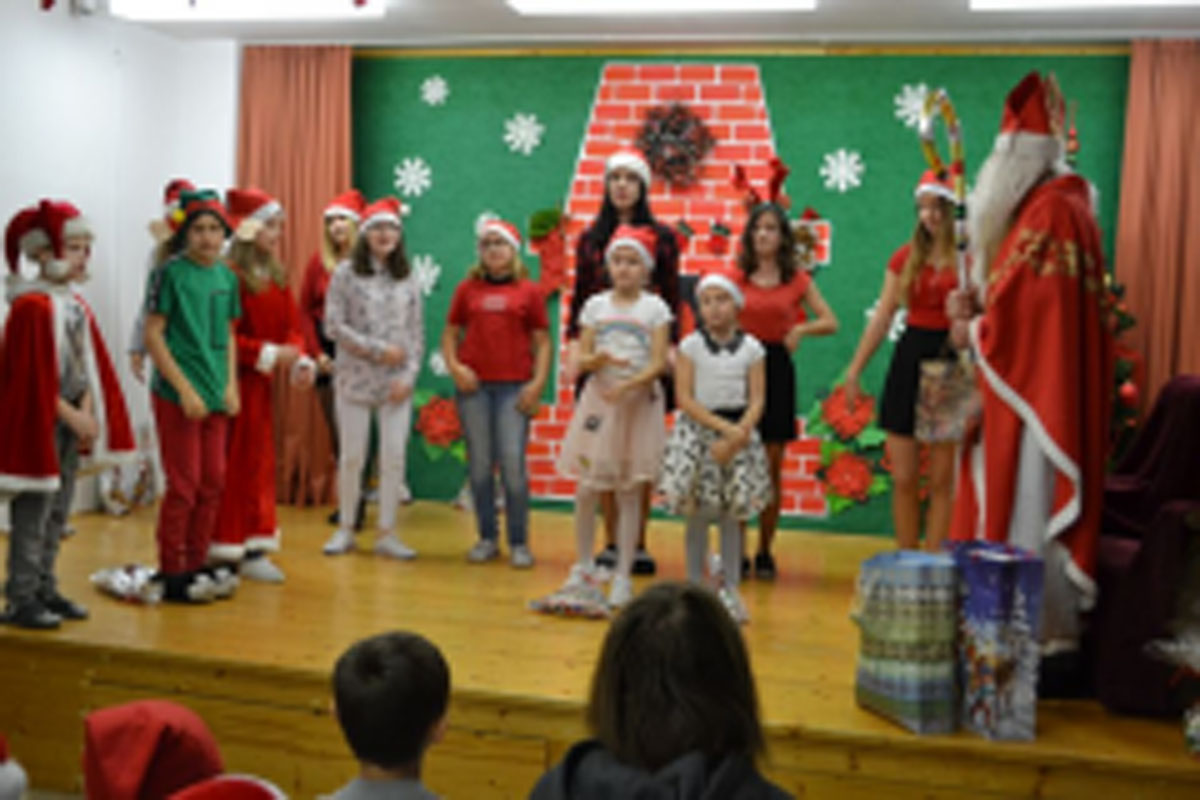 Uczniowie siedzą na krzesłach. Na głowach mają czerwone czapki Mikołaja. Oglądają występ na scenie. Mikołaj siedzi w fotelu na scenie i rozdaje prezenty.