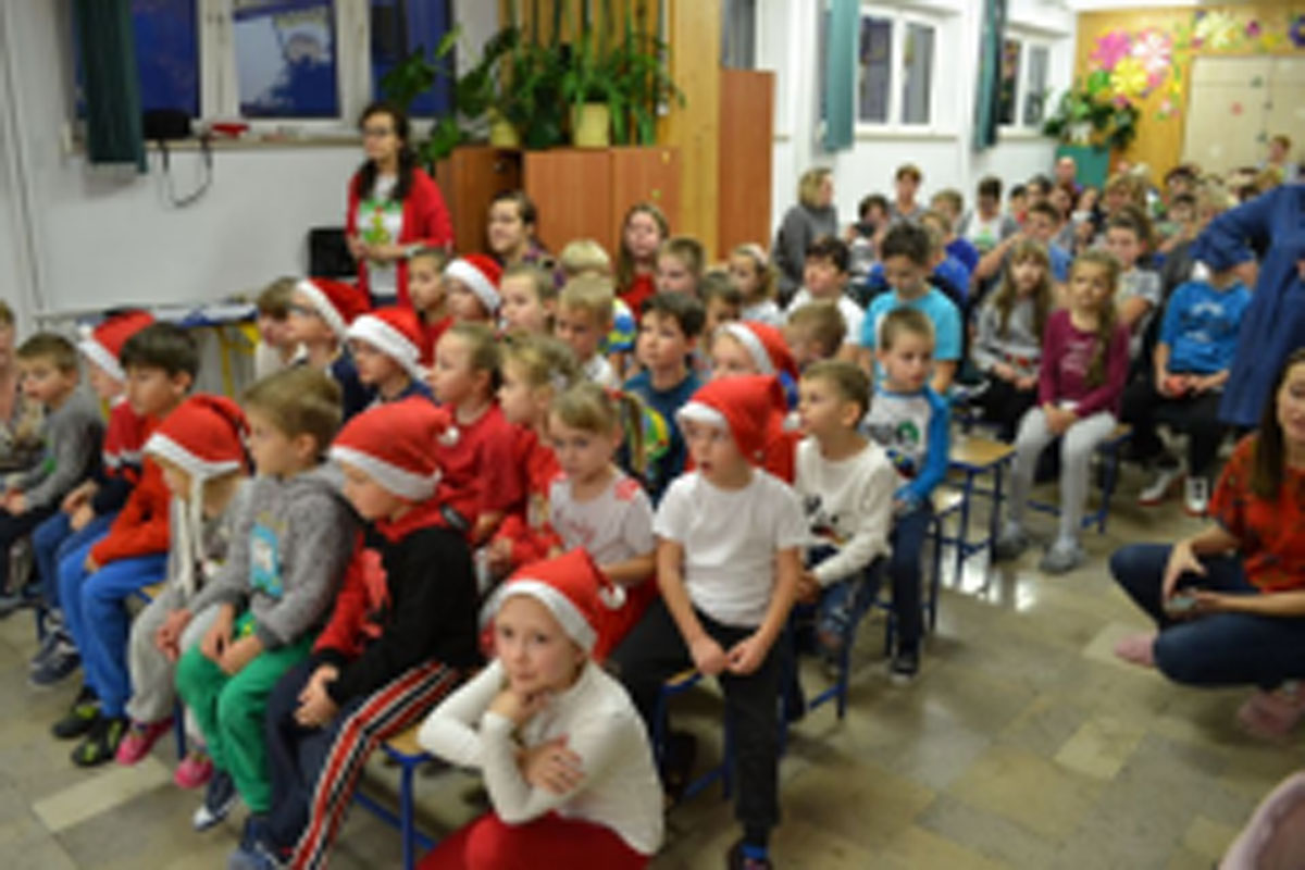 Uczniowie siedzą na krzesłach. Na głowach mają czerwone czapki Mikołaja. Oglądają występ na scenie. Mikołaj siedzi w fotelu na scenie i rozdaje prezenty.