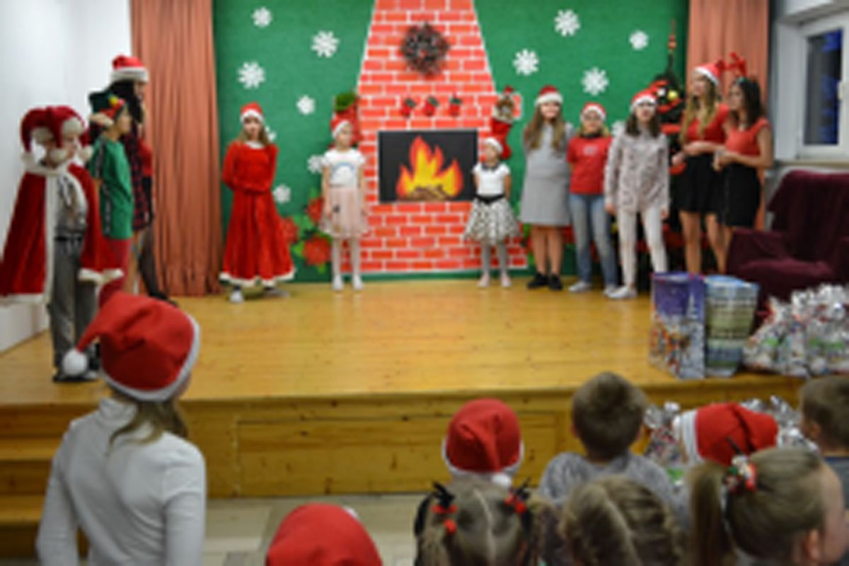 Uczniowie siedzą na krzesłach. Na głowach mają czerwone czapki Mikołaja. Oglądają występ na scenie. Mikołaj siedzi w fotelu na scenie i rozdaje prezenty.