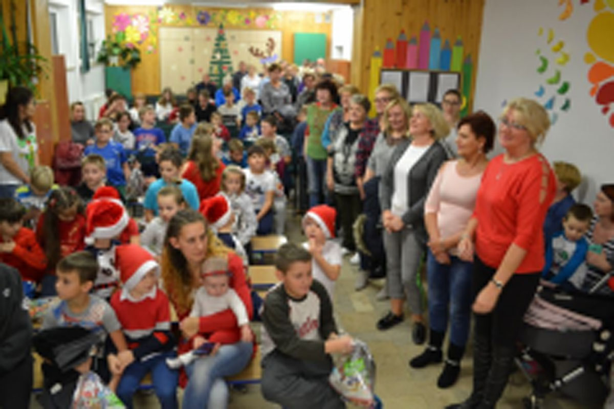 Uczniowie siedzą na krzesłach. Na głowach mają czerwone czapki Mikołaja. Oglądają występ na scenie. Mikołaj siedzi w fotelu na scenie i rozdaje prezenty.