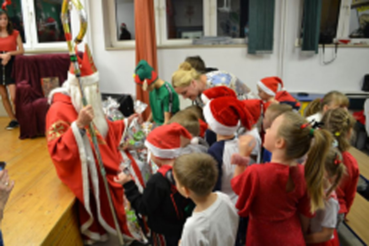 Uczniowie siedzą na krzesłach. Na głowach mają czerwone czapki Mikołaja. Oglądają występ na scenie. Mikołaj siedzi w fotelu na scenie i rozdaje prezenty.