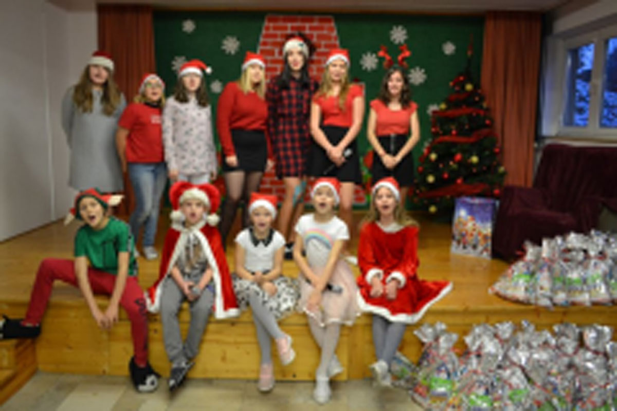 Uczniowie siedzą na krzesłach. Na głowach mają czerwone czapki Mikołaja. Oglądają występ na scenie. Mikołaj siedzi w fotelu na scenie i rozdaje prezenty.