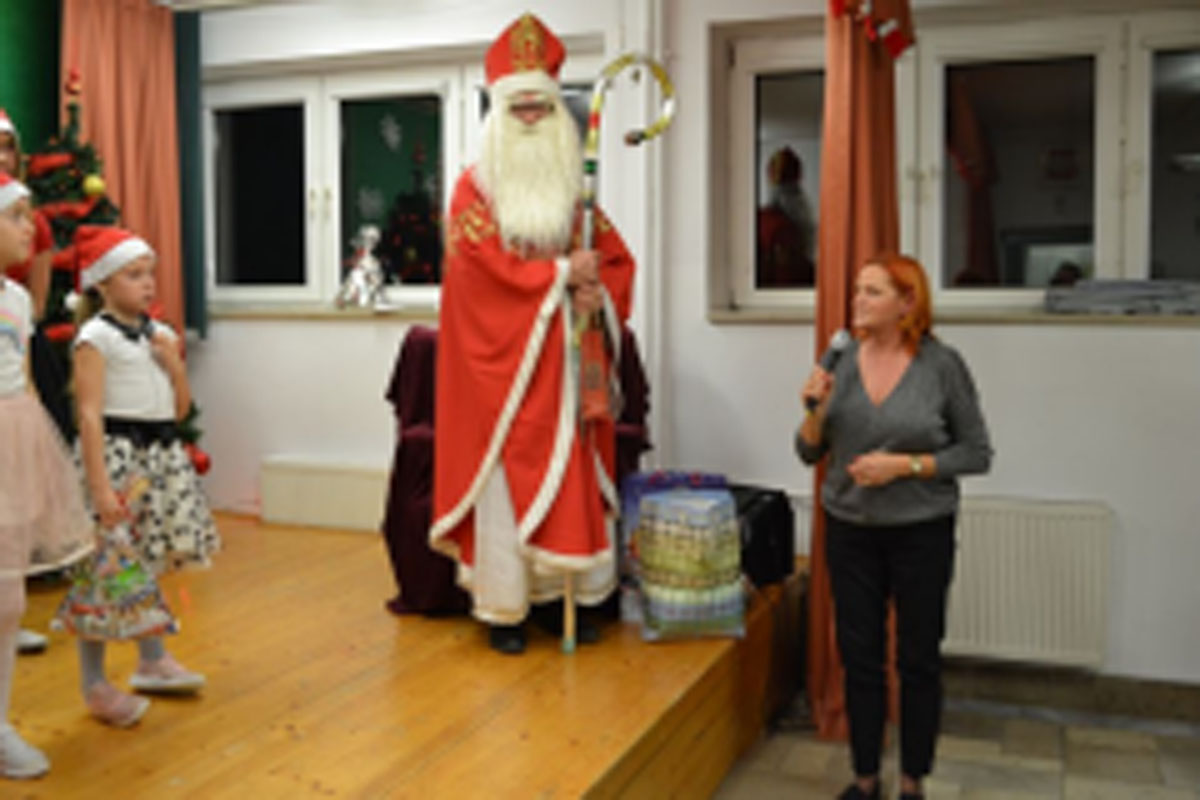 Uczniowie siedzą na krzesłach. Na głowach mają czerwone czapki Mikołaja. Oglądają występ na scenie. Mikołaj siedzi w fotelu na scenie i rozdaje prezenty.