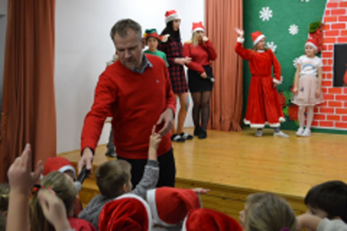 Uczniowie siedzą na krzesłach. Na głowach mają czerwone czapki Mikołaja. Oglądają występ na scenie. Mikołaj siedzi w fotelu na scenie i rozdaje prezenty.