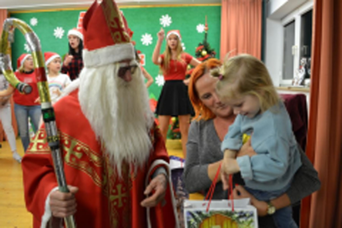 Uczniowie siedzą na krzesłach. Na głowach mają czerwone czapki Mikołaja. Oglądają występ na scenie. Mikołaj siedzi w fotelu na scenie i rozdaje prezenty.