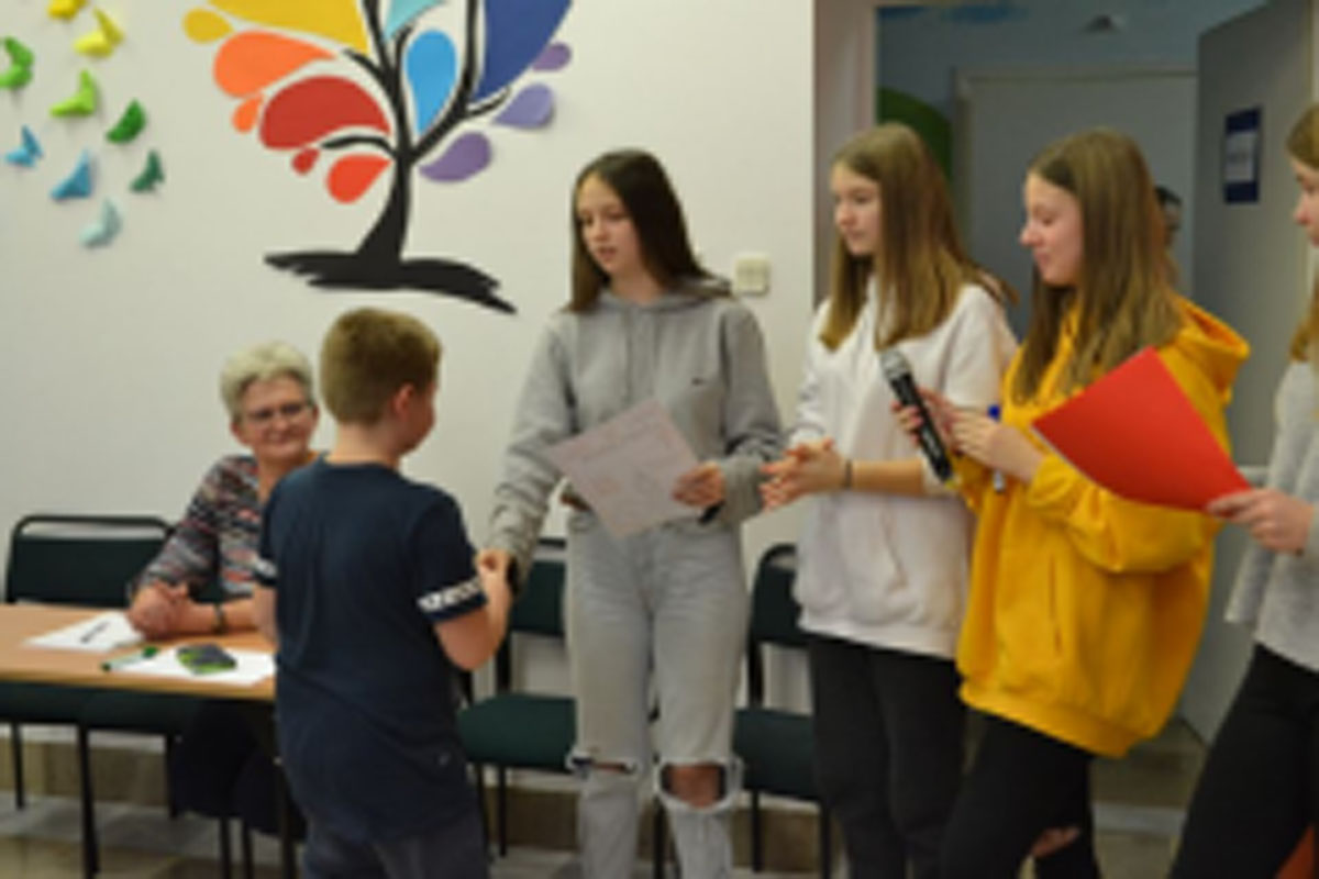 Uczniowie występują na scenie, trzymają mikrofony. Członkowie jury siedzą przy stoliku, wręczają dyplomy. Wspólne zdjęcie uczniów.