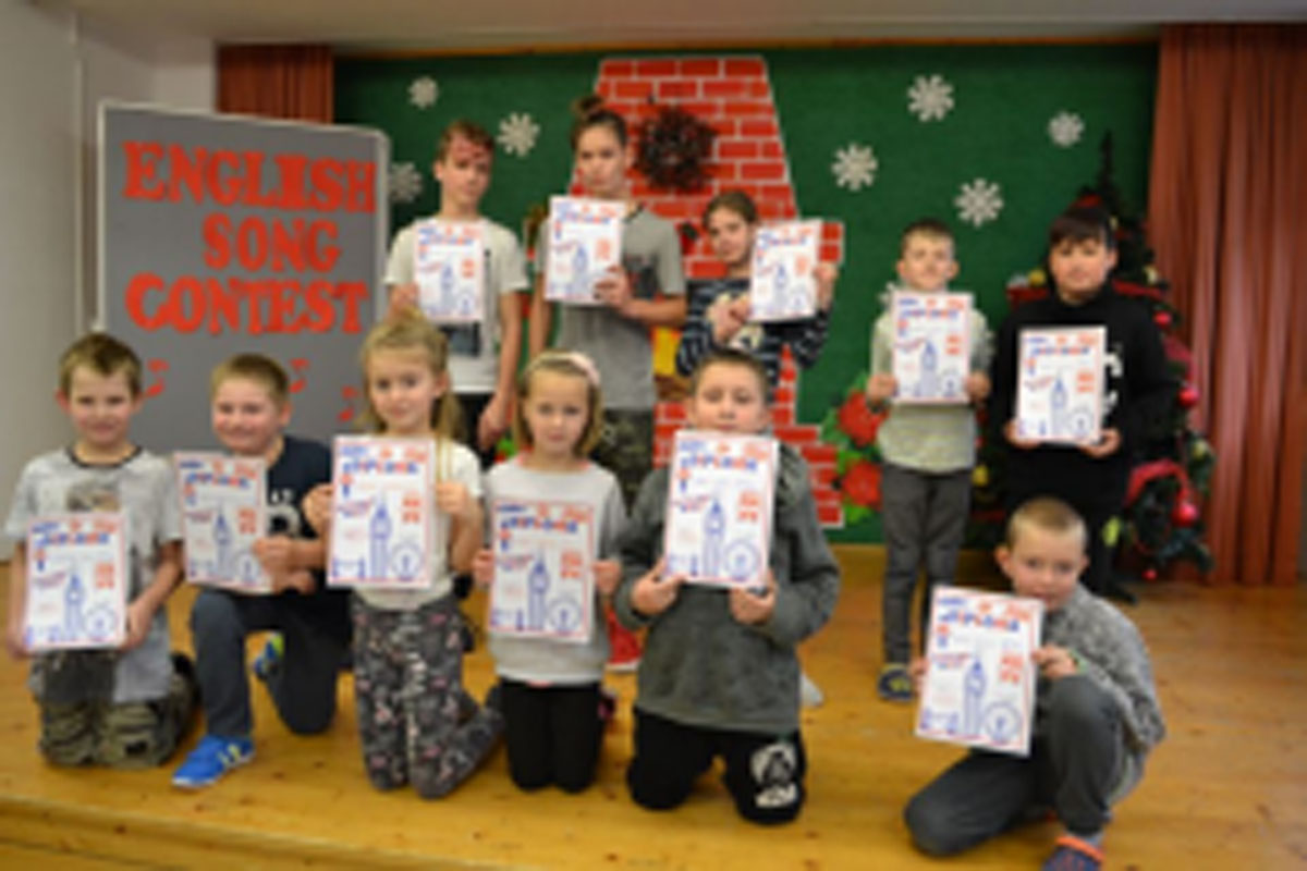 Uczniowie występują na scenie, trzymają mikrofony. Członkowie jury siedzą przy stoliku, wręczają dyplomy. Wspólne zdjęcie uczniów.