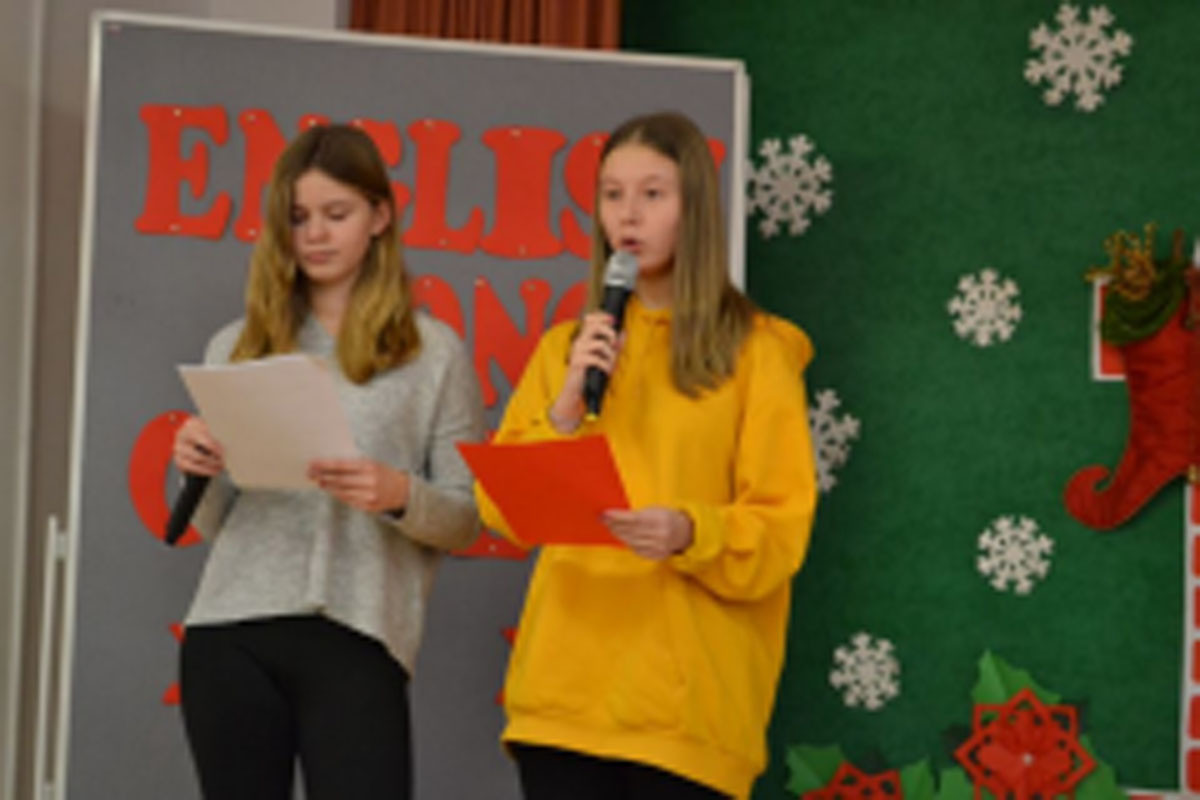 Uczniowie występują na scenie, trzymają mikrofony. Członkowie jury siedzą przy stoliku, wręczają dyplomy. Wspólne zdjęcie uczniów.