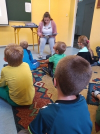 Dzieci w maseczkach siedzą na dywanie. Jedna starsza uczennica siedzi naprzeciwko dzieci i czyta im z dużej książki.