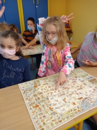 Dzieci w maseczkach siedzą na dywanie. Jedna starsza uczennica siedzi naprzeciwko dzieci i czyta im z dużej książki.