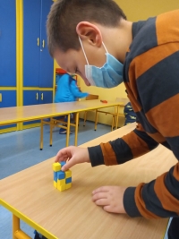 Dzieci w maseczkach siedzą na dywanie. Jedna starsza uczennica siedzi naprzeciwko dzieci i czyta im z dużej książki.