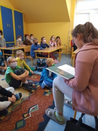 Dzieci w maseczkach siedzą na dywanie. Jedna starsza uczennica siedzi naprzeciwko dzieci i czyta im z dużej książki.