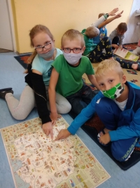 Dzieci w maseczkach siedzą na dywanie. Jedna starsza uczennica siedzi naprzeciwko dzieci i czyta im z dużej książki.