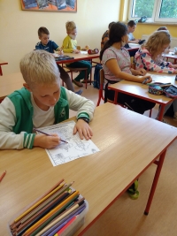 Dzieci w maseczkach siedzą na dywanie. Jedna starsza uczennica siedzi naprzeciwko dzieci i czyta im z dużej książki.