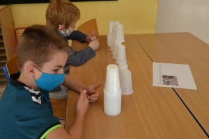 Uczniowie przy stolikach,  na których są kubki plastikowe ułożone w wieże, łyżeczki plastikowe w pojemniku i obok niego, gry planszowe i folia przeźroczysta z wynikami działań. Uczniowie siedzą na krzesłach patrzą na tablicę multimedialną.