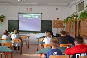 Uczniowie przy stolikach,  na których są kubki plastikowe ułożone w wieże, łyżeczki plastikowe w pojemniku i obok niego, gry planszowe i folia przeźroczysta z wynikami działań. Uczniowie siedzą na krzesłach patrzą na tablicę multimedialną.