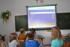 Uczniowie przy stolikach,  na których są kubki plastikowe ułożone w wieże, łyżeczki plastikowe w pojemniku i obok niego, gry planszowe i folia przeźroczysta z wynikami działań. Uczniowie siedzą na krzesłach patrzą na tablicę multimedialną.