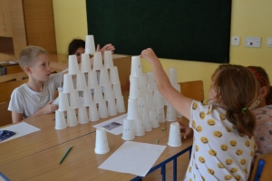 Uczniowie przy stolikach,  na których są kubki plastikowe ułożone w wieże, łyżeczki plastikowe w pojemniku i obok niego, gry planszowe i folia przeźroczysta z wynikami działań. Uczniowie siedzą na krzesłach patrzą na tablicę multimedialną.