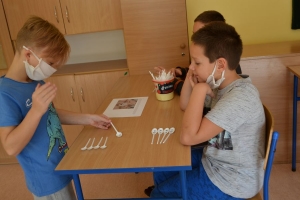 Uczniowie przy stolikach,  na których są kubki plastikowe ułożone w wieże, łyżeczki plastikowe w pojemniku i obok niego, gry planszowe i folia przeźroczysta z wynikami działań. Uczniowie siedzą na krzesłach patrzą na tablicę multimedialną.