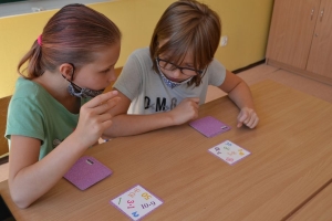 Uczniowie przy stolikach,  na których są kubki plastikowe ułożone w wieże, łyżeczki plastikowe w pojemniku i obok niego, gry planszowe i folia przeźroczysta z wynikami działań. Uczniowie siedzą na krzesłach patrzą na tablicę multimedialną.