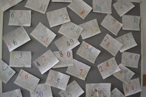 Uczniowie przy stolikach,  na których są kubki plastikowe ułożone w wieże, łyżeczki plastikowe w pojemniku i obok niego, gry planszowe i folia przeźroczysta z wynikami działań. Uczniowie siedzą na krzesłach patrzą na tablicę multimedialną.