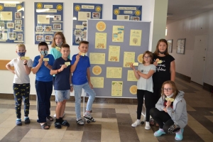 Uczniowie przy stolikach,  na których są kubki plastikowe ułożone w wieże, łyżeczki plastikowe w pojemniku i obok niego, gry planszowe i folia przeźroczysta z wynikami działań. Uczniowie siedzą na krzesłach patrzą na tablicę multimedialną.