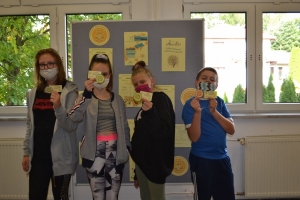 Uczniowie przy stolikach,  na których są kubki plastikowe ułożone w wieże, łyżeczki plastikowe w pojemniku i obok niego, gry planszowe i folia przeźroczysta z wynikami działań. Uczniowie siedzą na krzesłach patrzą na tablicę multimedialną.