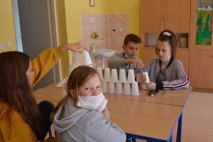 Uczniowie przy stolikach,  na których są kubki plastikowe ułożone w wieże, łyżeczki plastikowe w pojemniku i obok niego, gry planszowe i folia przeźroczysta z wynikami działań. Uczniowie siedzą na krzesłach patrzą na tablicę multimedialną.