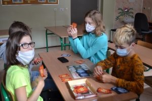 Uczniowie przy stolikach,  na których są kubki plastikowe ułożone w wieże, łyżeczki plastikowe w pojemniku i obok niego, gry planszowe i folia przeźroczysta z wynikami działań. Uczniowie siedzą na krzesłach patrzą na tablicę multimedialną.