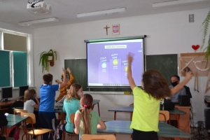 Uczniowie przy stolikach,  na których są kubki plastikowe ułożone w wieże, łyżeczki plastikowe w pojemniku i obok niego, gry planszowe i folia przeźroczysta z wynikami działań. Uczniowie siedzą na krzesłach patrzą na tablicę multimedialną.