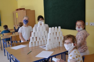 Uczniowie przy stolikach,  na których są kubki plastikowe ułożone w wieże, łyżeczki plastikowe w pojemniku i obok niego, gry planszowe i folia przeźroczysta z wynikami działań. Uczniowie siedzą na krzesłach patrzą na tablicę multimedialną.