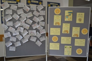 Uczniowie przy stolikach,  na których są kubki plastikowe ułożone w wieże, łyżeczki plastikowe w pojemniku i obok niego, gry planszowe i folia przeźroczysta z wynikami działań. Uczniowie siedzą na krzesłach patrzą na tablicę multimedialną.