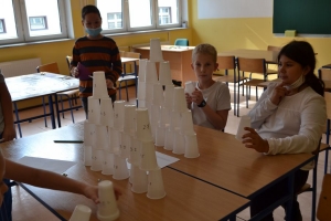 Uczniowie przy stolikach,  na których są kubki plastikowe ułożone w wieże, łyżeczki plastikowe w pojemniku i obok niego, gry planszowe i folia przeźroczysta z wynikami działań. Uczniowie siedzą na krzesłach patrzą na tablicę multimedialną.