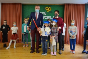 Uczniowie na scenie mówią do mikrofonu. Dyrektor mianuje pierwszoklasistów dużym ołówkiem.