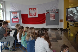 Na tle tablicy z biało-czerwonym sercem i napisem Serce dla Polski widoczna jest grupka dzieci w maseczkach. Jeden z chłopów ma uniesioną prawa rękę z palcami ułożonymi w znak zwycięstwa.ZSU Rabka