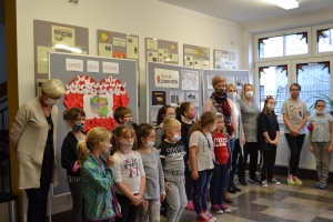 Na tle tablicy z biało-czerwonym sercem i napisem Serce dla Polski widoczna jest grupka dzieci w maseczkach. Jeden z chłopów ma uniesioną prawa rękę z palcami ułożonymi w znak zwycięstwa.SU Rabka