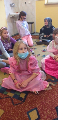 Grupa dzieci, przebranych w stroje bajkowych postaci  siedzą na dywanie i słuchają czytanej im bajki.