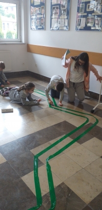 Na podłodze wyklejona jest trasa. Dzieci po kolei, pstrykają w swoją nakrętkę, tak aby przesunąć ją do przodu i by nie wypaść z trasy.