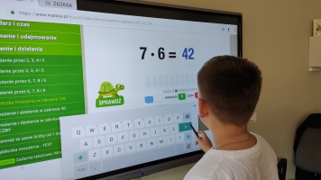 Uczniowie przy komputerach i stolikach. Układają wierze z plastikowych kubków, losują łyżeczki z zadaniami, grają w gry planszowe, rozwiązują zadania w programie komputerowym.