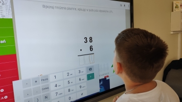 Uczniowie przy komputerach i stolikach. Układają wierze z plastikowych kubków, losują łyżeczki z zadaniami, grają w gry planszowe, rozwiązują zadania w programie komputerowym.