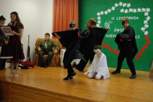 Uczniowie na scenie śpiewają pieśni. Koło sceny poczet sztandarowy, na scenie uczniowie przebrani w czarne stroje zaborców i  na biało za orła. Uczniowie składają znicze pod krzyżem.