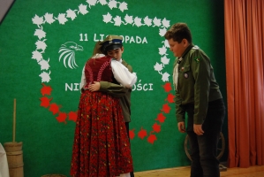 Uczniowie na scenie śpiewają pieśni. Koło sceny poczet sztandarowy, na scenie uczniowie przebrani w czarne stroje zaborców i  na biało za orła. Uczniowie składają znicze pod krzyżem.