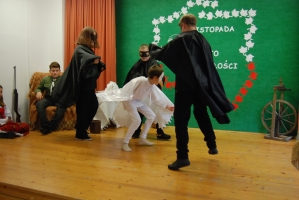 Uczniowie na scenie śpiewają pieśni. Koło sceny poczet sztandarowy, na scenie uczniowie przebrani w czarne stroje zaborców i  na biało za orła. Uczniowie składają znicze pod krzyżem.